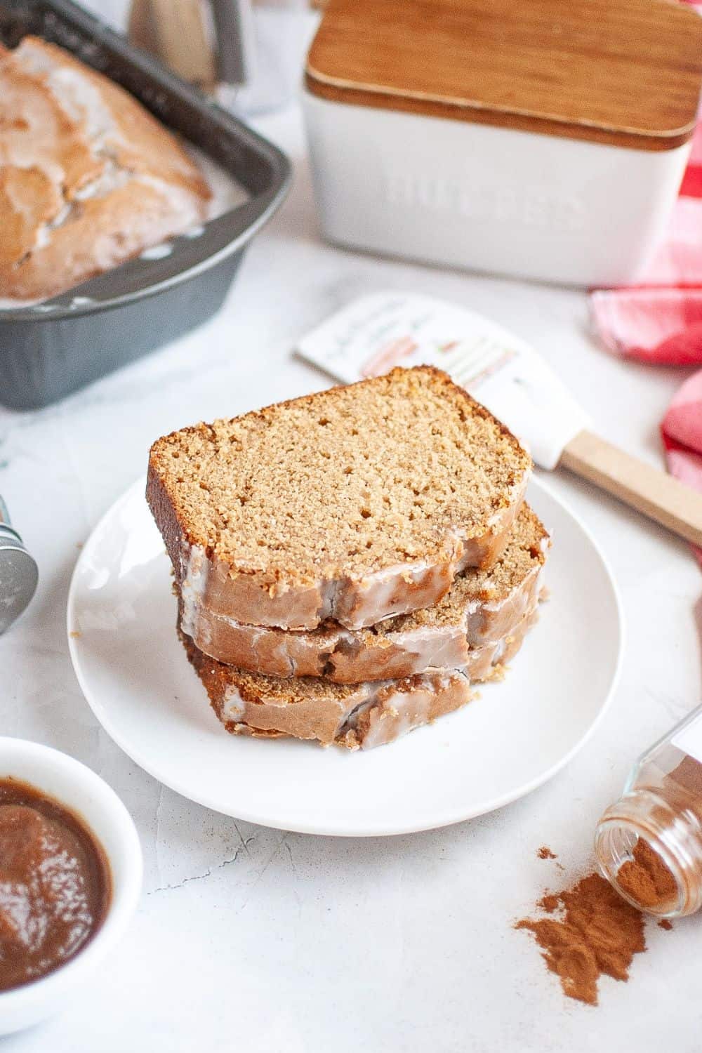 Slices of quickbread made with apple butter served on a white plate, with various ingredients around the plate. The loaf pan of apple butter bread is in the background.