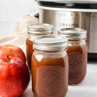 Three jars of slow cooker apple butter in front of a Crock Pot. Two fresh apples are next to the jars.