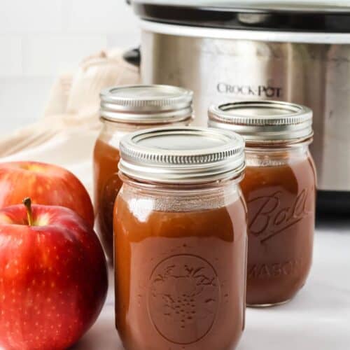 Three jars of slow cooker apple butter in front of a Crock Pot. Two fresh apples are next to the jars.