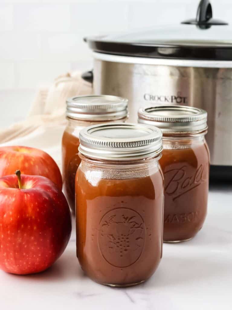 Three jars of slow cooker apple butter in front of a Crock Pot. Two fresh apples are next to the jars.