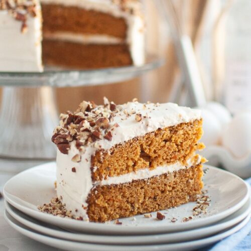 Slice of canned carrot cake served on a white plate, with the remainder of the cake on a cake stand in the background.