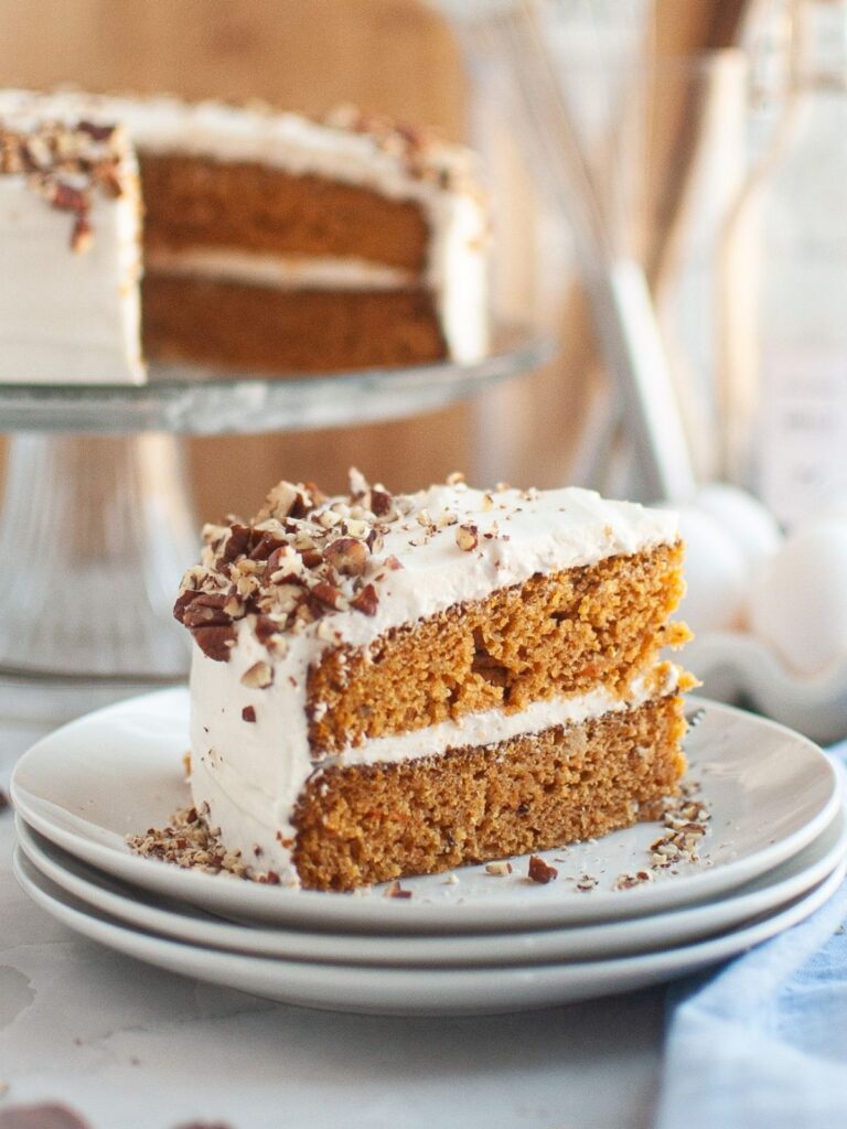 Slice of canned carrot cake served on a white plate, with the remainder of the cake on a cake stand in the background.