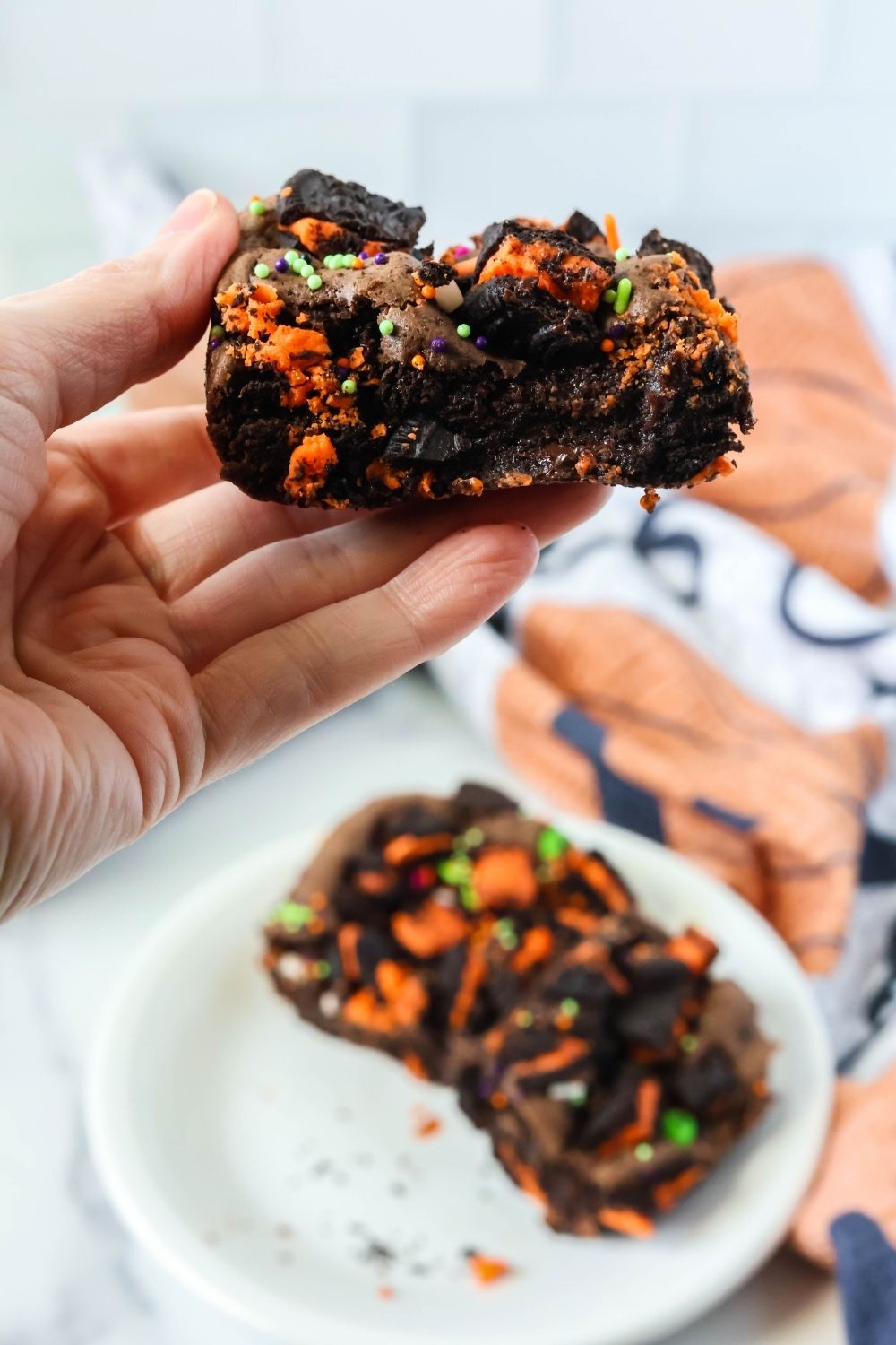 A woman's hand holds a chocolate Oreo cake bar made for Halloween, complete with orange Oreo filling, plus green, orange, and purple Halloween sprinkles.