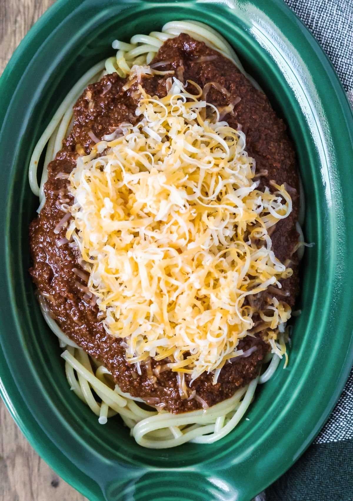 Overhead view of a green dish filled with spaghetti, Cincinnati chili made from scratch, and shredded cheese.