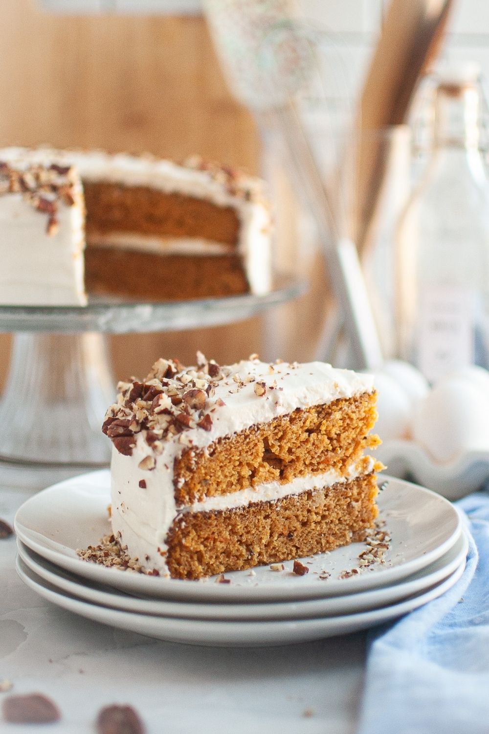 Side view of a slice of cake made with canned carrots and topped with cream cheese frosting. The remainder of the layer cake is on a cake stand in the background.