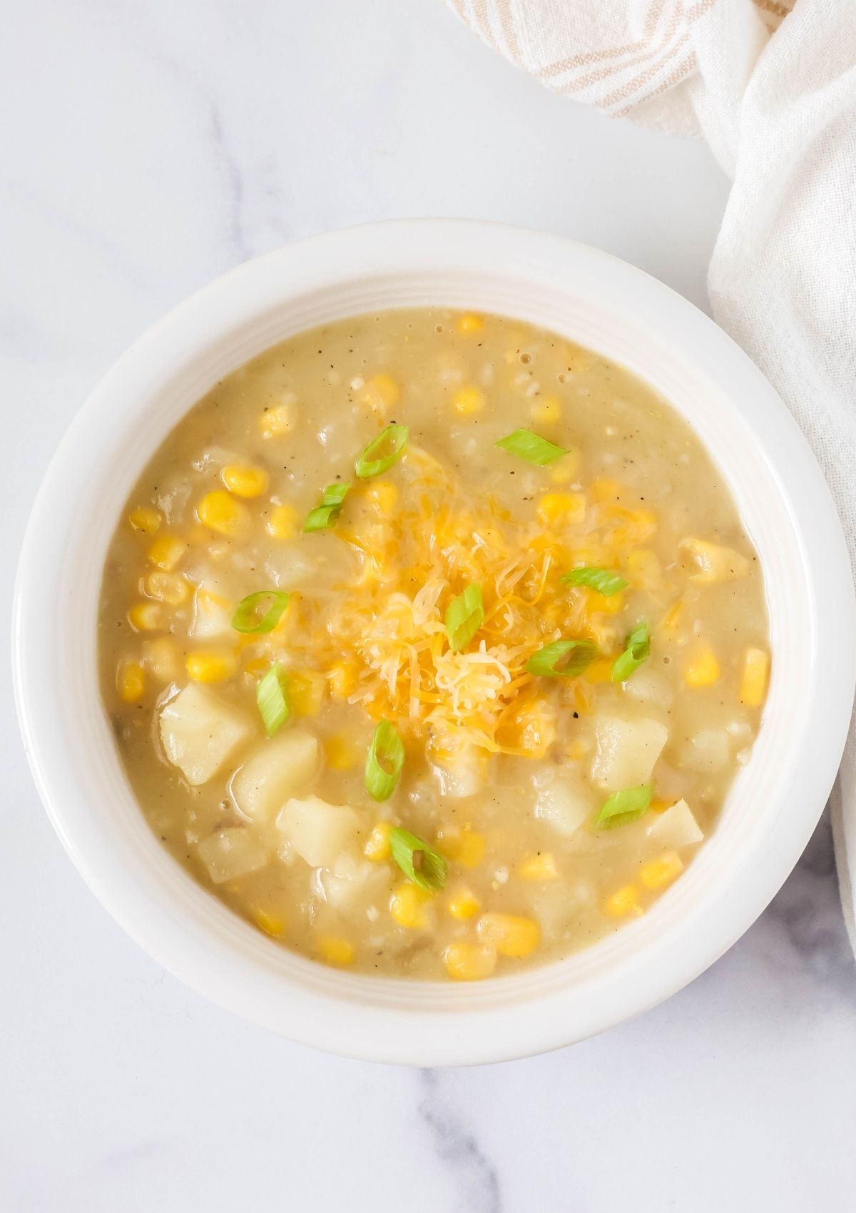 Overhead view of a white bowl filled with easy potato corn chowder soup, garnished with shredded cheese and sliced green onions.