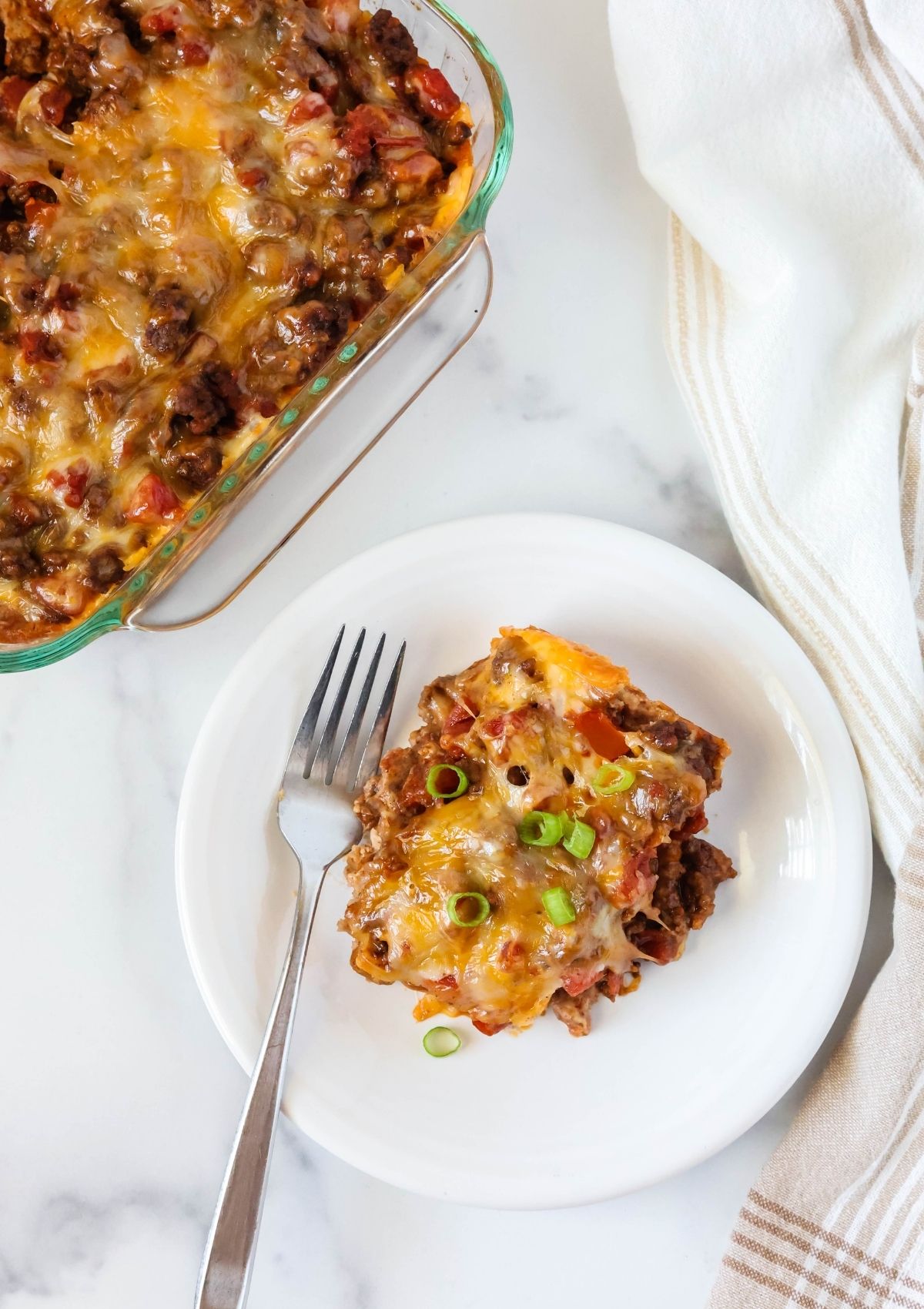 Overhead view of a white plate with a serving of easy taco casserole on it, alongside a fork. The casserole dish is in the background.