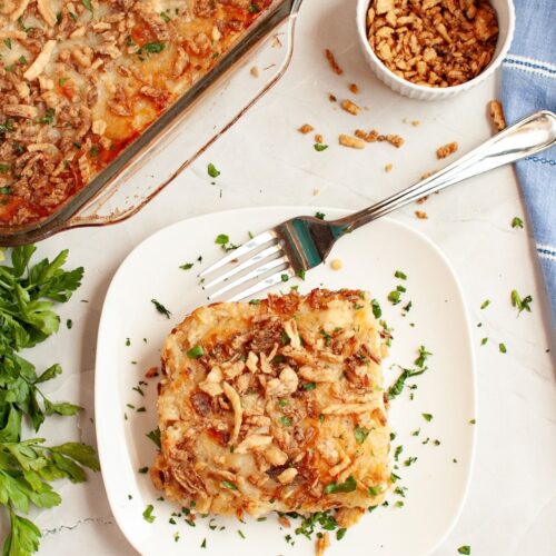 Serving of french onion chicken and rice on a white plate with a fork next to it. The casserole dish is in the background, along with some crispy fried onions.
