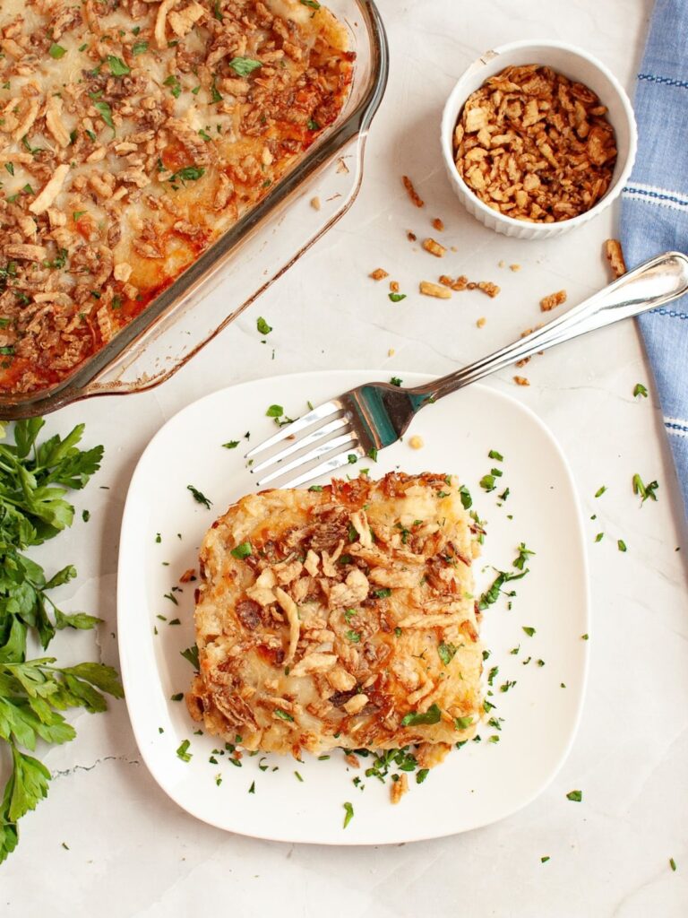 Serving of french onion chicken and rice on a white plate with a fork next to it. The casserole dish is in the background, along with some crispy fried onions.