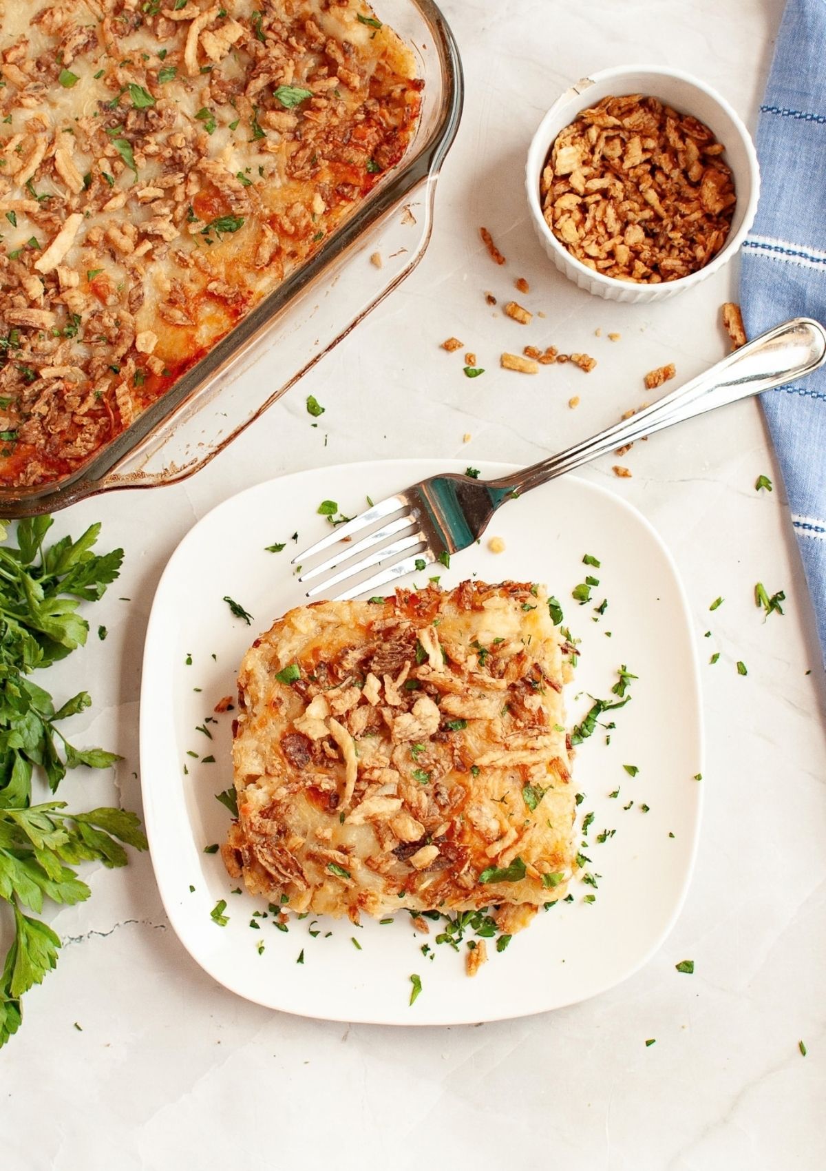 A helping of french onion rice and chicken casserole served on a white plate. A fork is on the plate, and the glass baking dish is next to the plate, along with crispy fried onions.