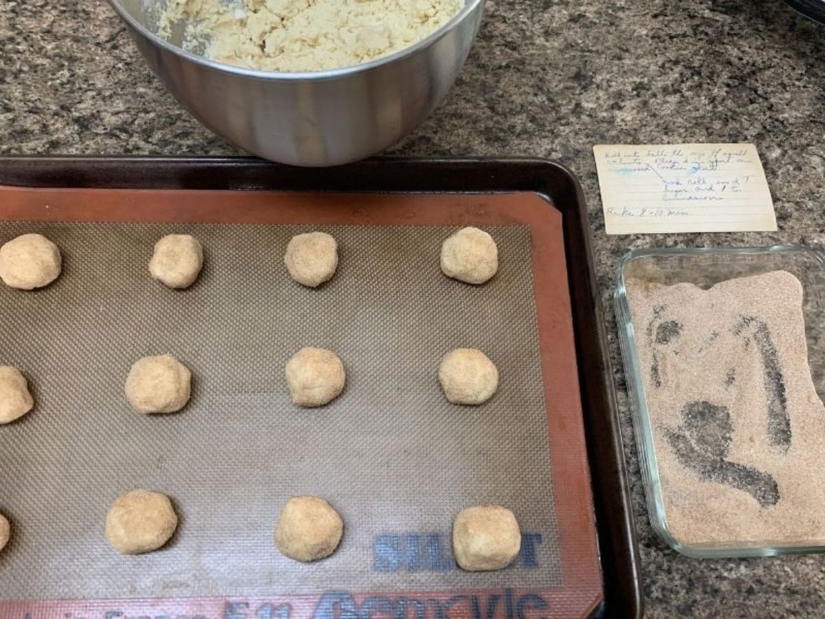 Snickerdoodle cookie dough balls on a baking sheet, with cinnamon sugar next to the baking sheet, as well as grandma's recipe card.