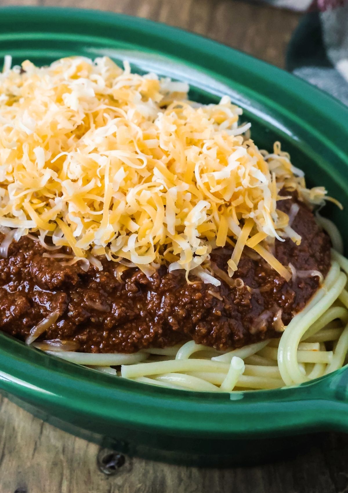 Close-up view of homemade copycat Skyline chili served over a bed of spaghetti noodles and topped with shredded cheese, served in a green dish.
