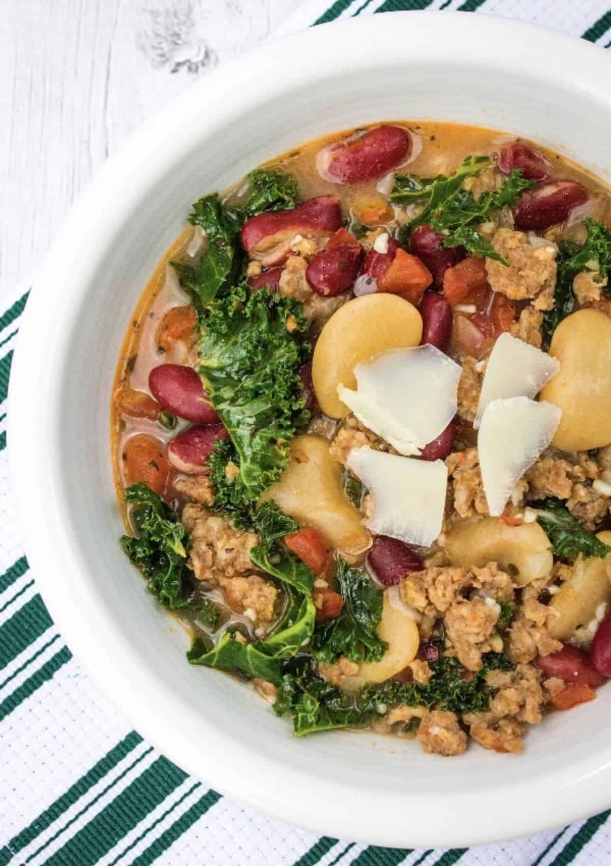 Overhead view of a white bowl filled with sausage and bean soup, garnished with parmesan shavings.