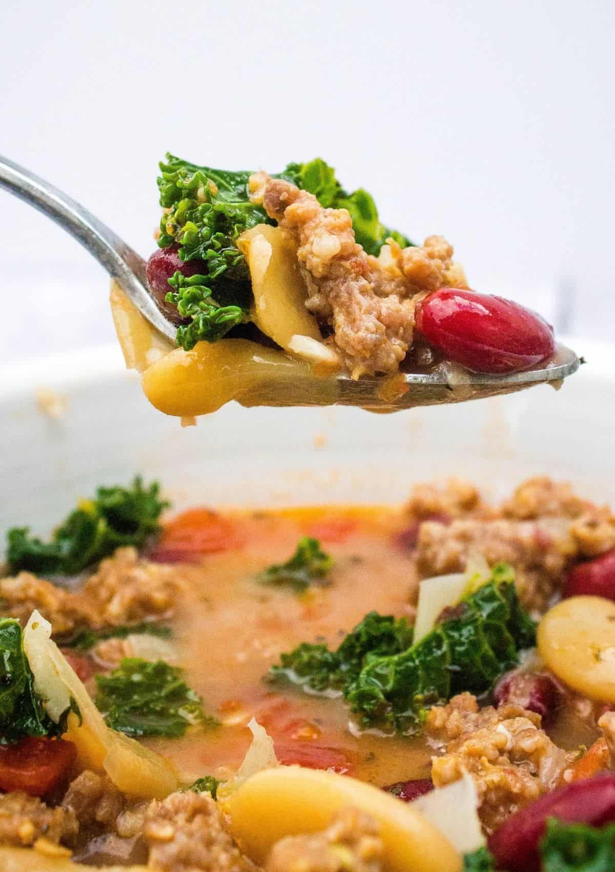 a spoon lifts a bite of bean and italian sausage soup from a white bowl.
