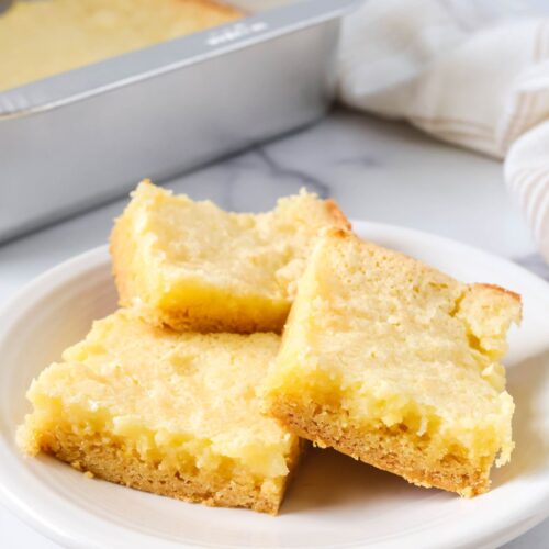 Ooey gooey butter cake squares served on a white plate, with the pan of remaining bars in the background.
