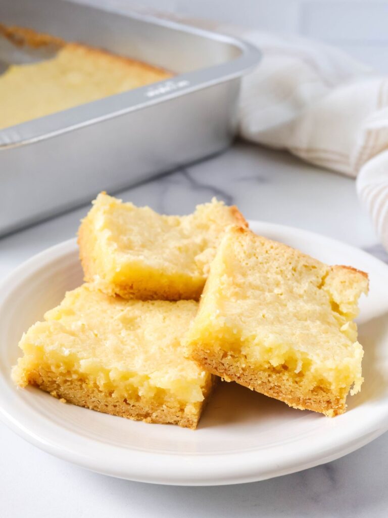 Ooey gooey butter cake squares served on a white plate, with the pan of remaining bars in the background.
