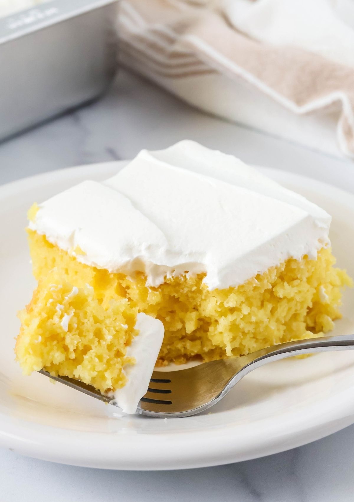 Slice of pineapple cake made with lemon cake mix, topped with Cool Whip, on a white plate. A fork has cut a bite out of the cake.