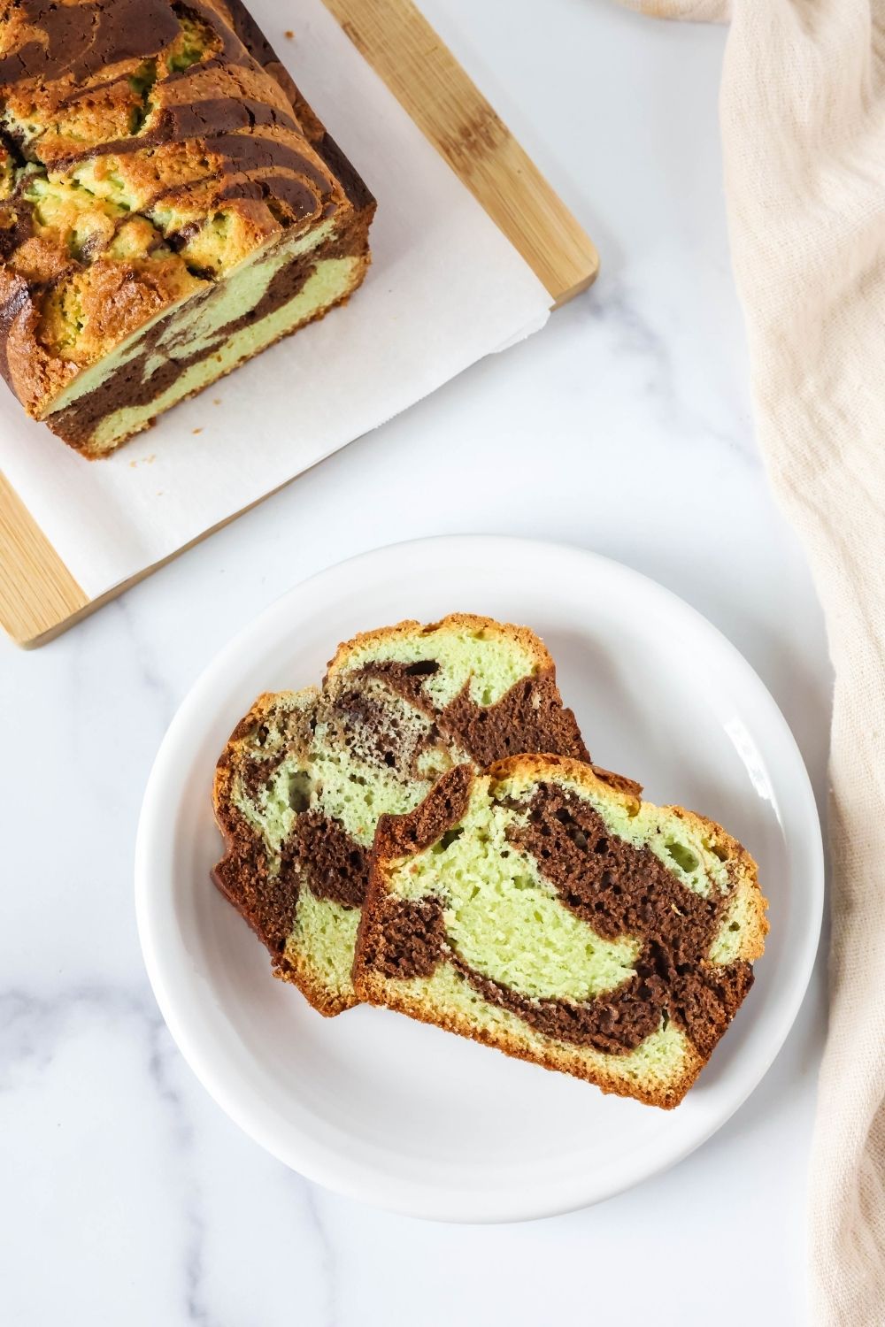 Overhead view of two slices of pistachio chocolate loaf on a white plate, with the remainder of the loaf of quick bread on a parchment-lined cutting board.