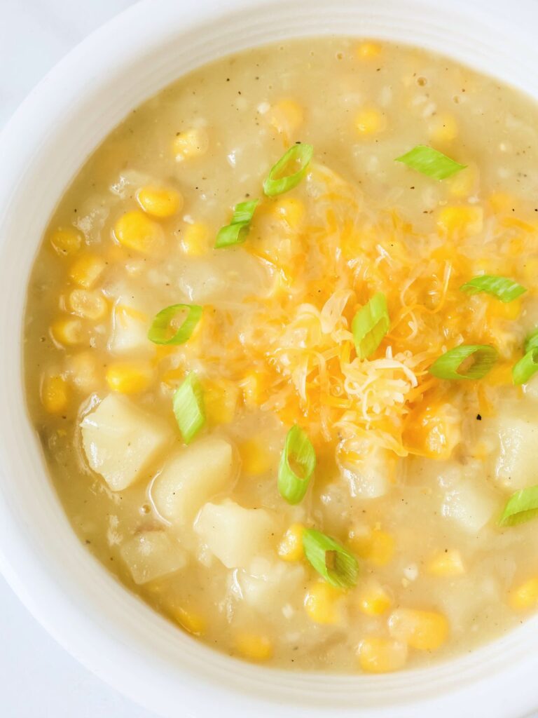 Potato corn chowder served in a white bowl, garnished with shredded cheese and sliced green onions.