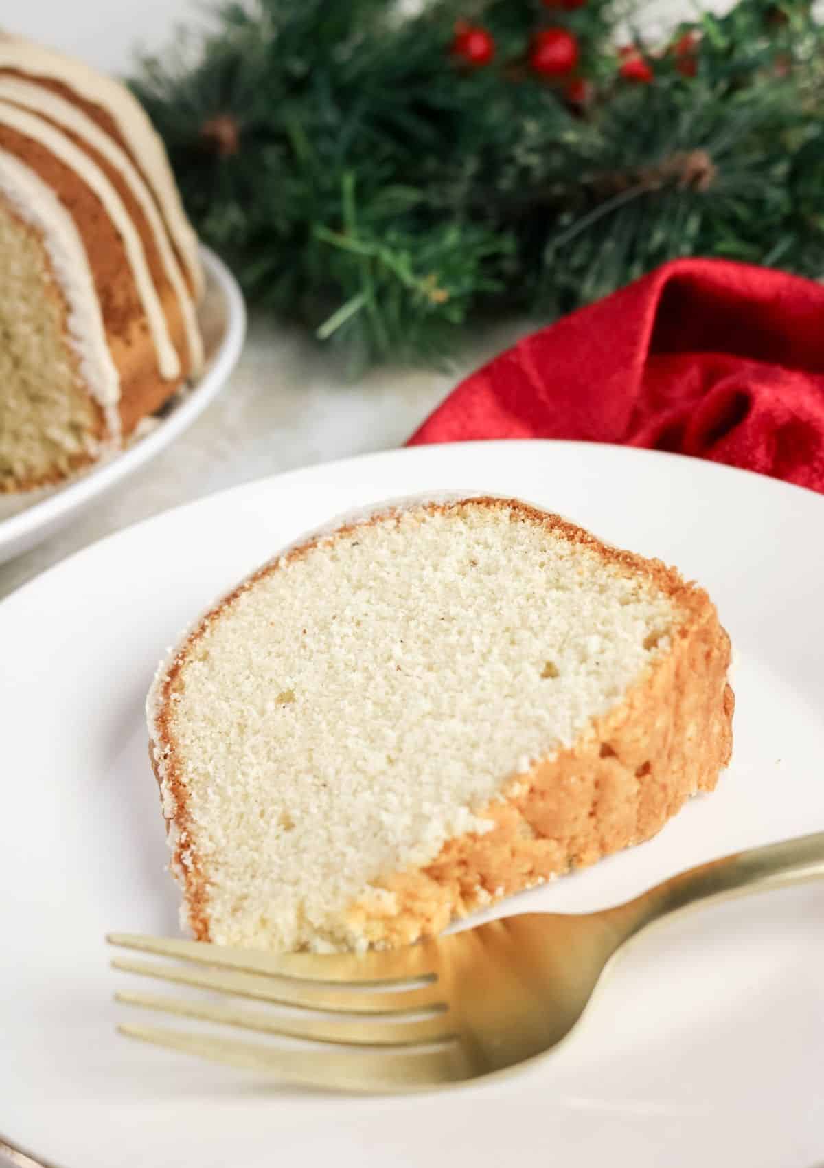 A slice of eggnog pound cake served on a white plate with a gold fork. The rest of the bundt cake is in the backround, along with some holiday decor.
