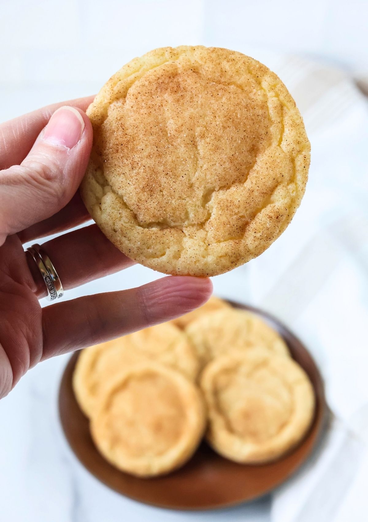 A hand holds a soft and chewy snickerdoodle cookie. A plate of more cookies is in the background.