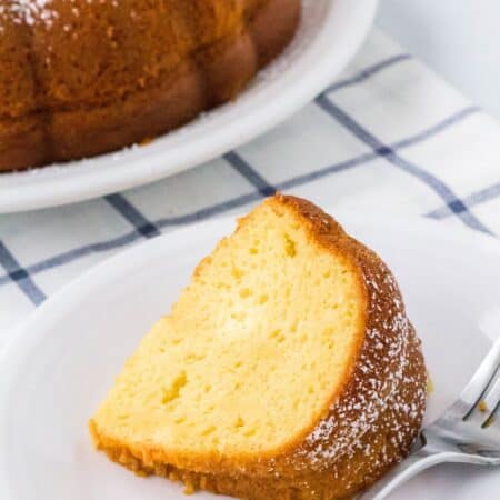 slice of vanilla pudding bundt cake on a white plate, with the remainder of the bundt cake in the background