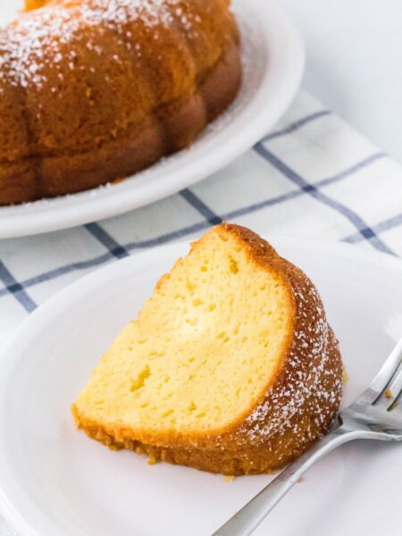 slice of vanilla pudding bundt cake on a white plate, with the remainder of the bundt cake in the background