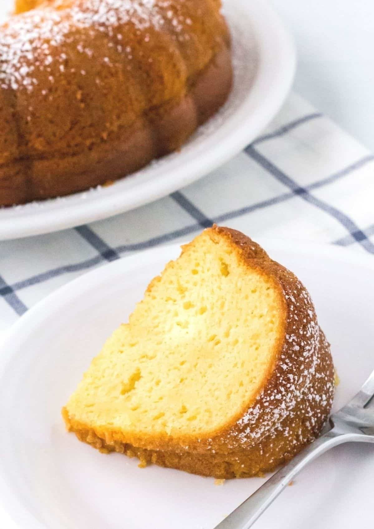 slice of vanilla pudding cake lying on its side on a white plate, with a fork next to it and the remainder of the bundt cake in the background