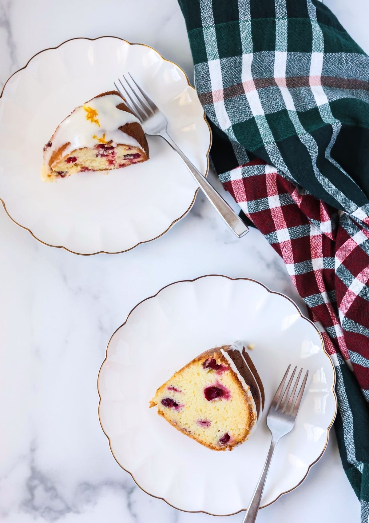Overhead view of two white scalloped dessert plates, each with a slice of cake mix cranberry pound cake on it. A green and red plaid kitchen towel is next to the plates.