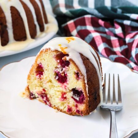 Slice of cranberry orange pound cake made from a cake mix served on a white dessert plate with a fork. A green and red kitchen towel is in the background, along with the remainder of the bundt cake.
