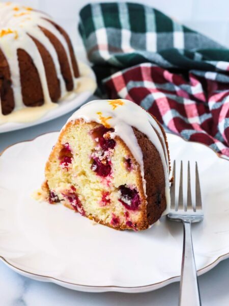 Slice of cranberry orange pound cake made from a cake mix served on a white dessert plate with a fork. A green and red kitchen towel is in the background, along with the remainder of the bundt cake.