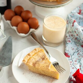 Slice of eggnog custard pie served on a white plate with a fork. The remainder of the whole pie, a glass of eggnog, and eggs are in the background.
