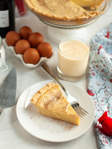 Slice of eggnog custard pie served on a white plate with a fork. The remainder of the whole pie, a glass of eggnog, and eggs are in the background.