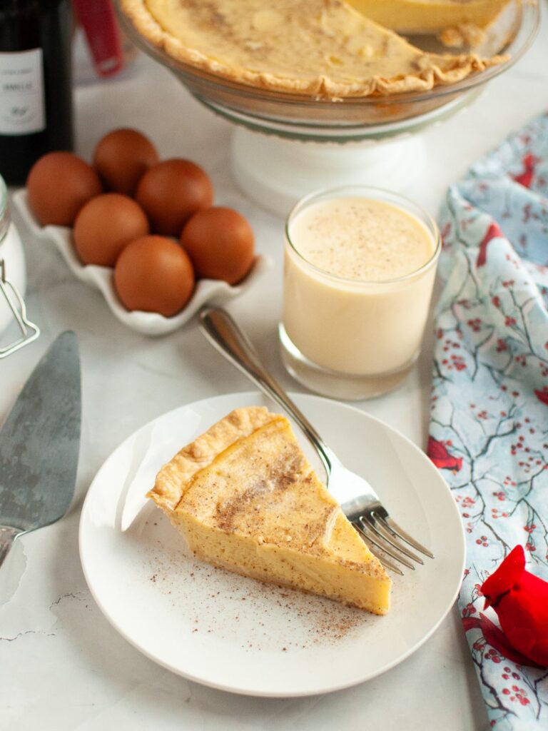 Slice of eggnog custard pie served on a white plate with a fork. The remainder of the whole pie, a glass of eggnog, and eggs are in the background.