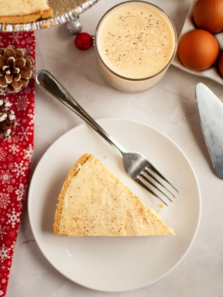 A slice of eggnog ice cream pie on a white plate with a fork. A glass of eggnog is next to the plate.