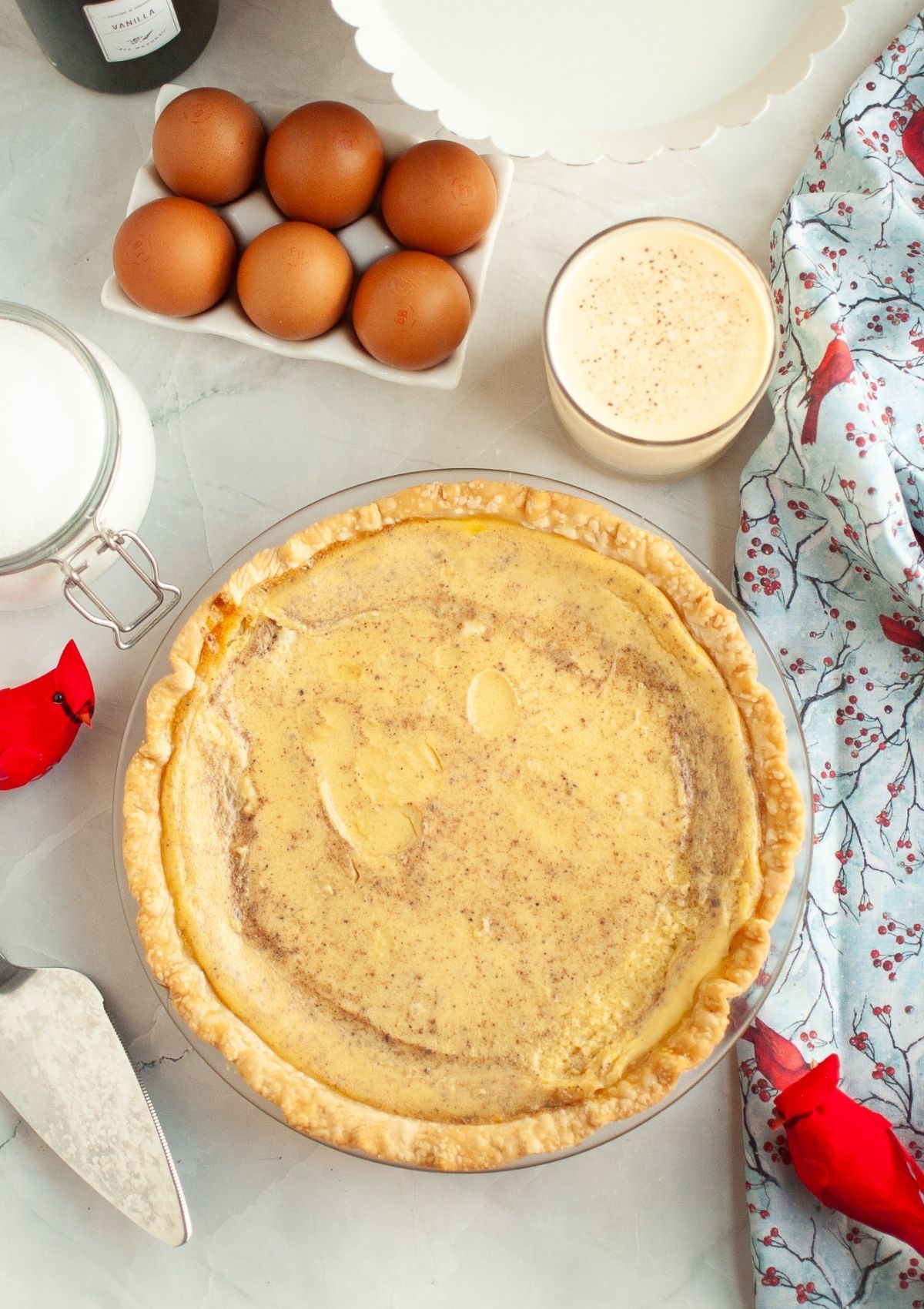 Overhead view of a holiday pie made with eggnog, in a pastry pie crust. The pie is surrounded by pie ingredients, such as eggnog, eggs, and sugar.