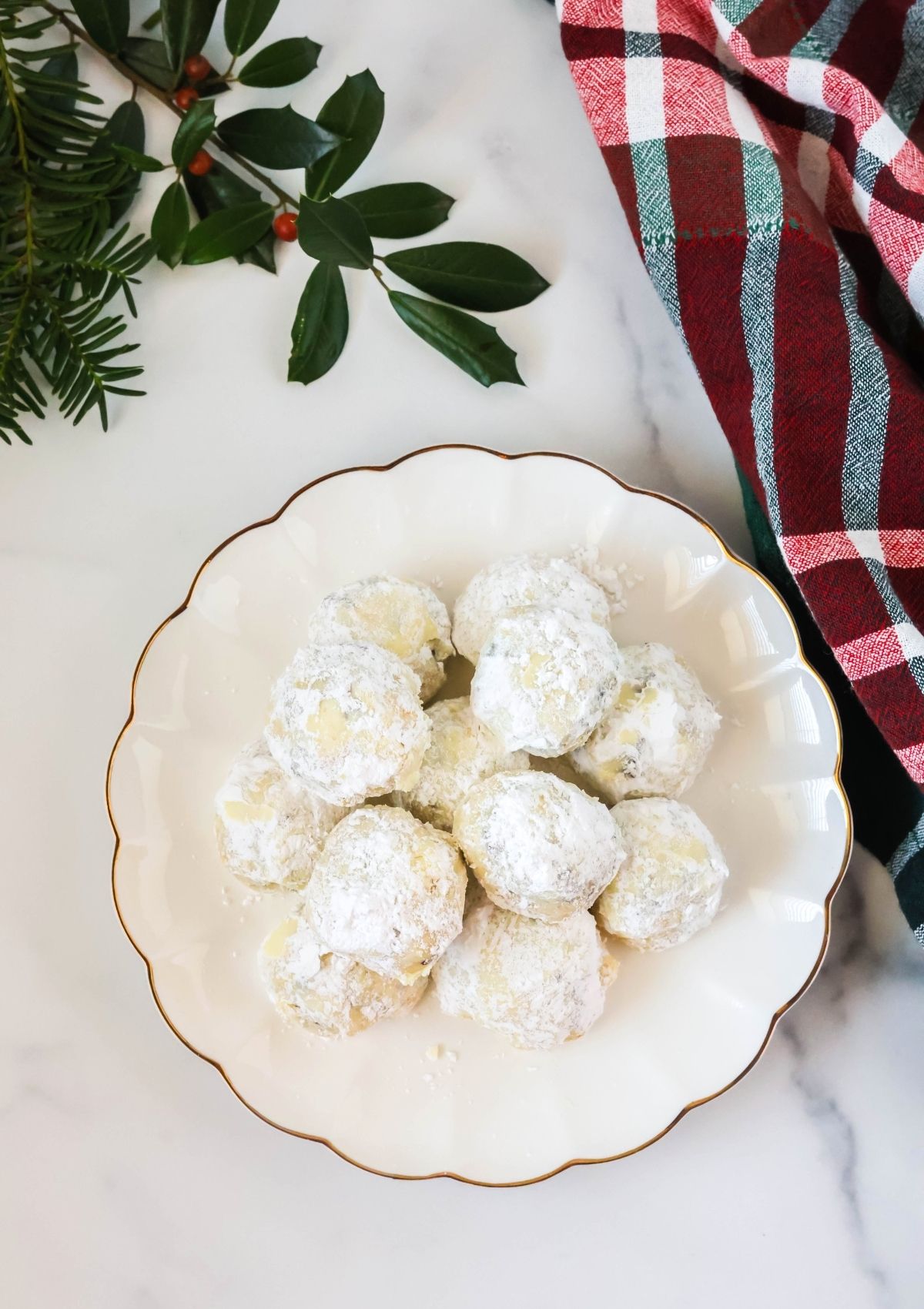 Overhead view of orange cranberry snowball cookies on a white plate, with festive greenery and a Christmas kitchen towel in the background.