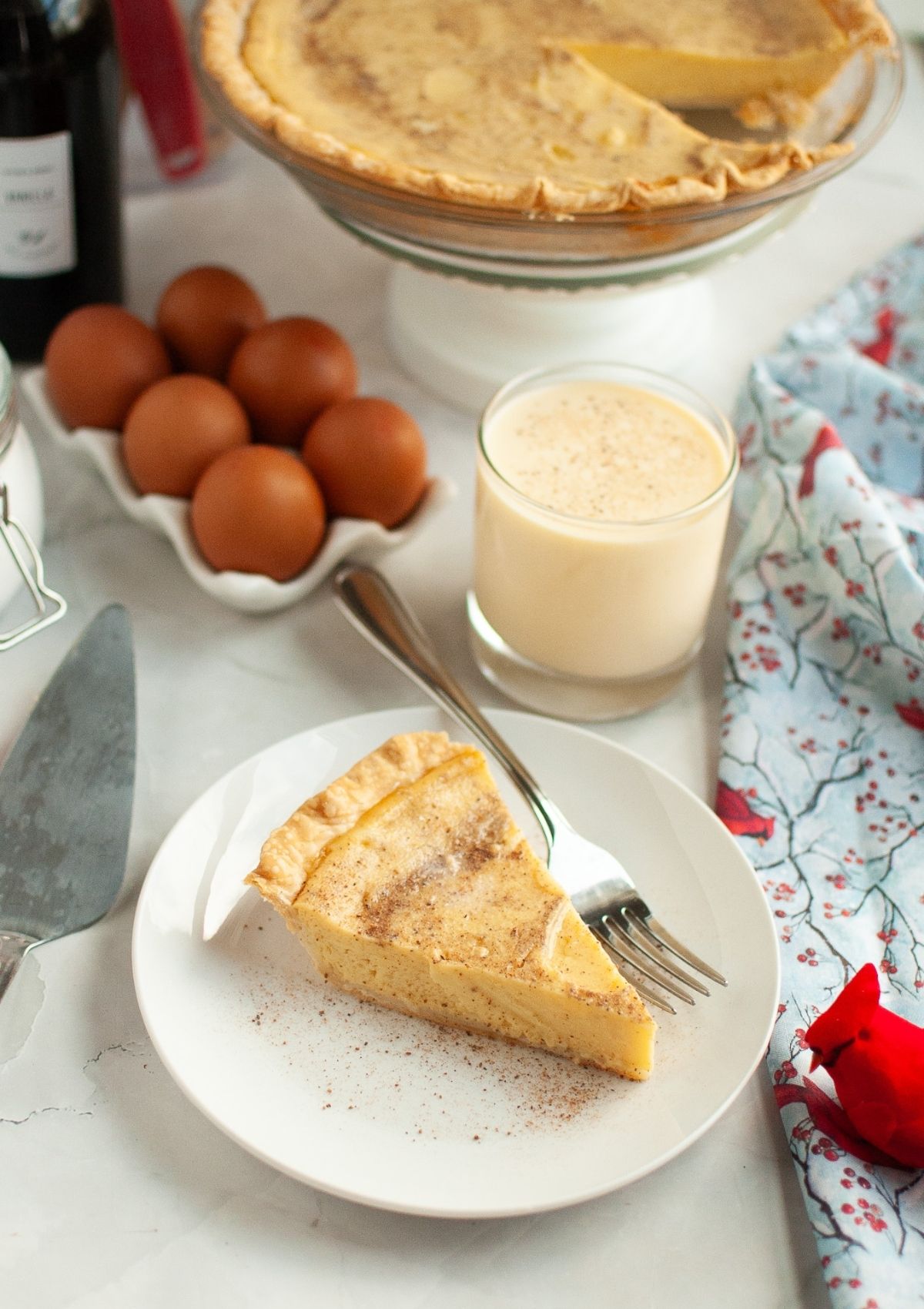 Slice of eggnog custard pie on a white plate with a fork. The remainder of the pie is in the background, along with a glass of eggnog and some eggs.