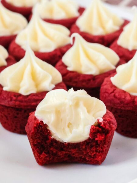Red velvet cookie cups served on a white plate. One of the cookie cups has had a bite taken out of it, showcasing the tender cookie crust and frosting filling.