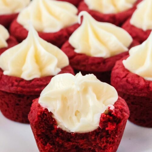 Red velvet cookie cups served on a white plate. One of the cookie cups has had a bite taken out of it, showcasing the tender cookie crust and frosting filling.