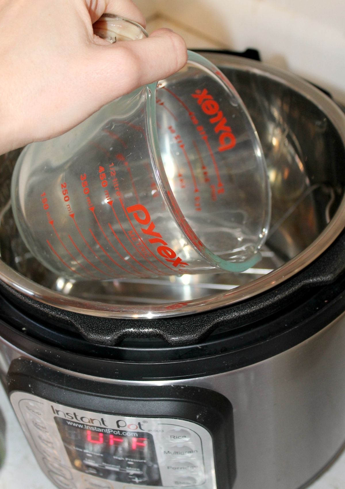 A woman's hand holds a glass measuring cup to pour a cup of water into the Instant Pot.